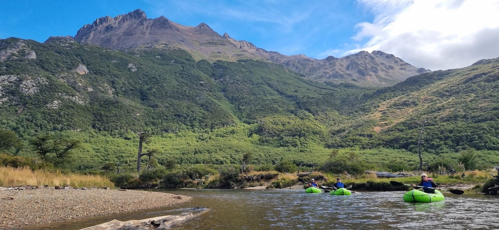 Experiencia de packrafting con XPLOR Turismo en Ushuaia, Argentina. Tres remeros en kayaks inflables verdes en un río con montañas y bosques de Tierra del Fuego al fondo.