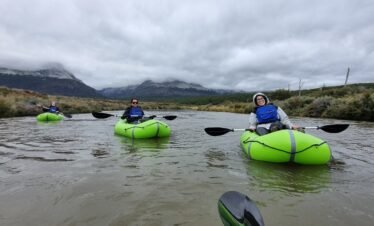 Tres turistas sonrientes remando en packrafts de color verde brillante por un río en Ushuaia, con las montañas de la Cordillera de los Andes cubiertas por nubes bajas al fondo. Excursión de aventura con XPLOR Turismo.