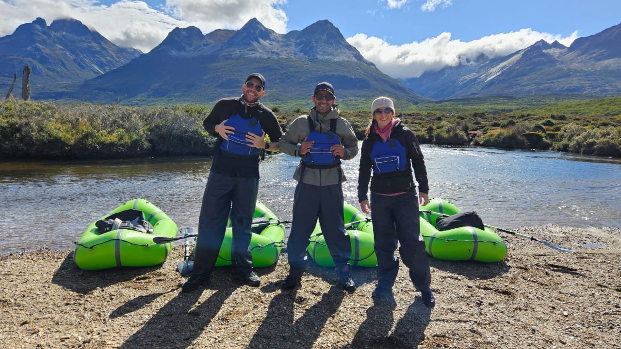 Tres turistas sonrientes equipados con chalecos salvavidas azules, posando junto a tres packrafts verdes en la orilla de un río en Ushuaia, con los picos nevados de los Andes Fueguinos de fondo. Excursión de aventura con XPLOR Turismo.
