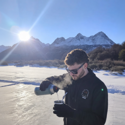 Aventurero sirviendo agua caliente en un mate frente a una laguna congelada y montañas nevadas al sol en Ushuaia, durante una excursión con Vértice Expediciones.