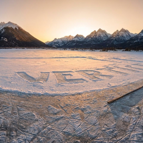 Palabra Vértice esculpida en la nieve sobre una laguna congelada al atardecer en Ushuaia, con marcas de patines en el hielo, un palo de hockey y la cordillera de fondo.